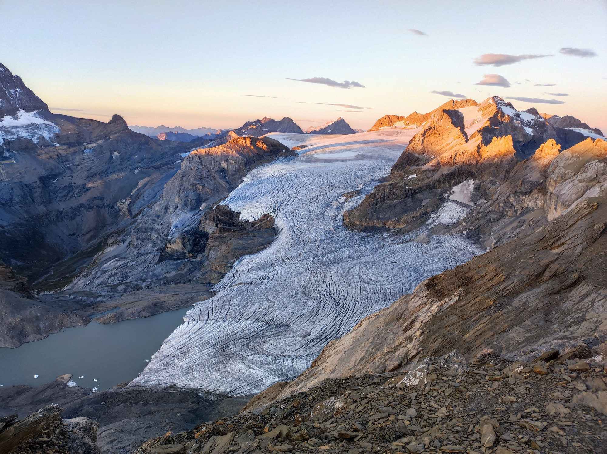 Snowy glacier winds through jagged mountains bathed in warm sunset light, with orange peaks and blue ice lining the valley.
