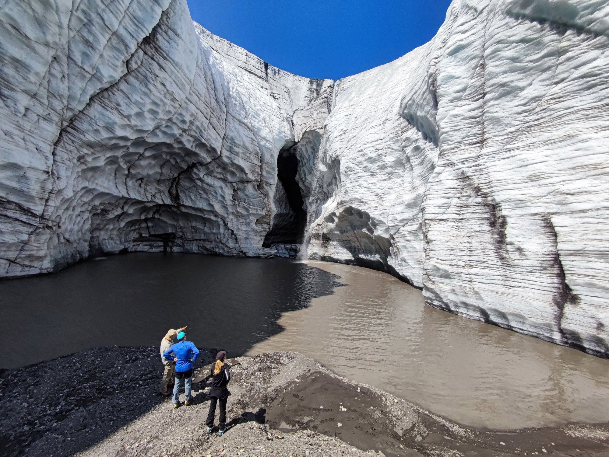 Group of four people standing on a rocky shore, facing a dark cave opening in a towering glacier wall under a clear blue sky