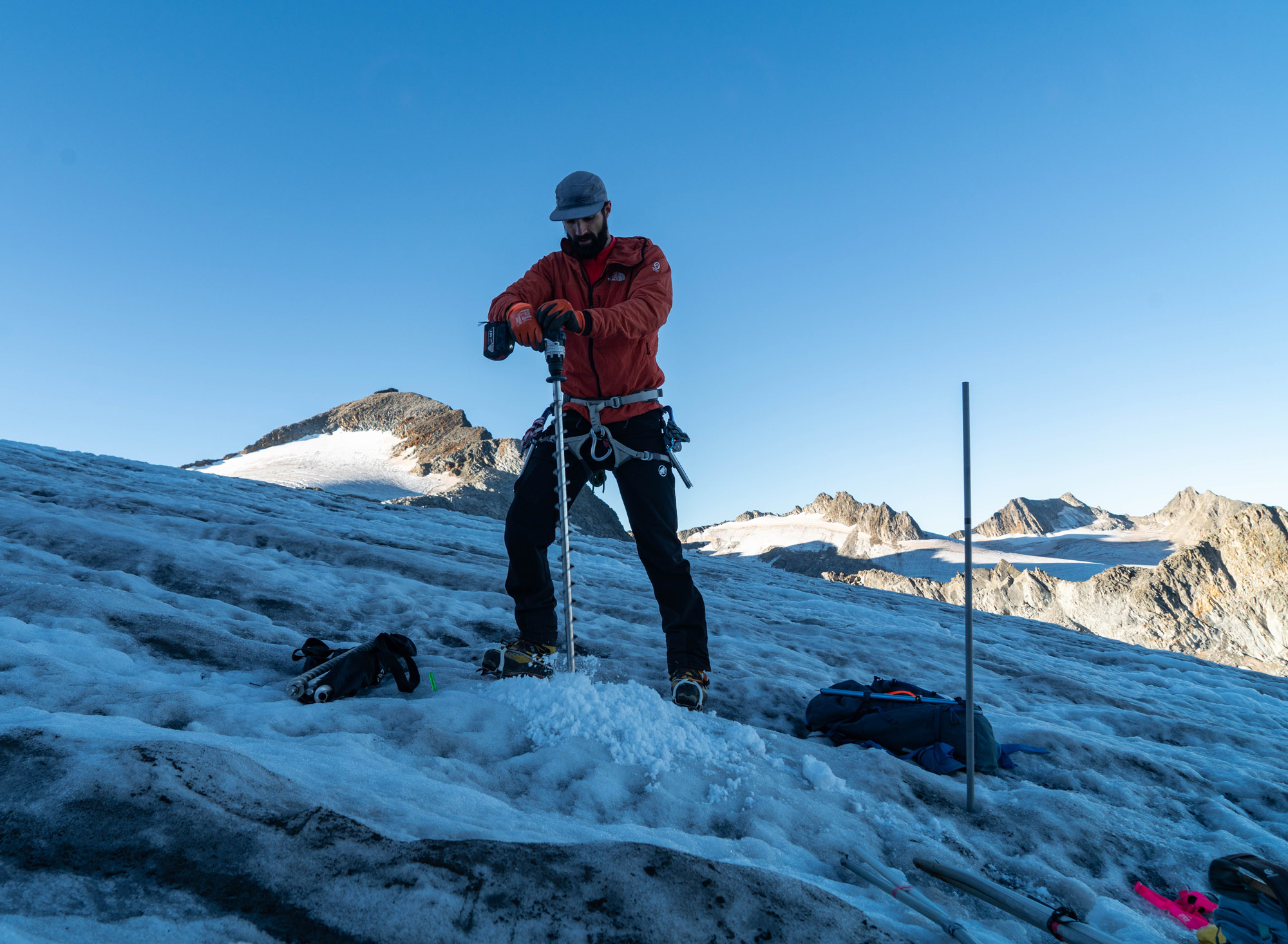 Mountaineer in a red jacket adjusting an ice axe on a snowy alpine slope under a clear blue sky.