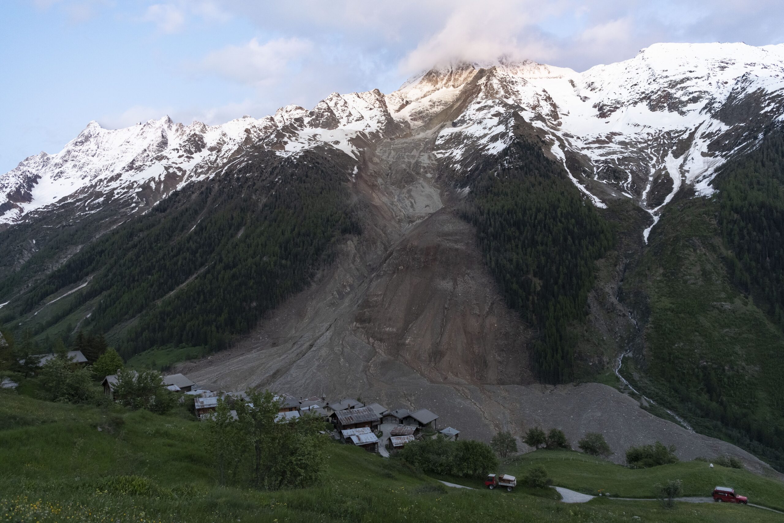 Snowy mountain range with jagged peaks above a green valley and a small village at the base.