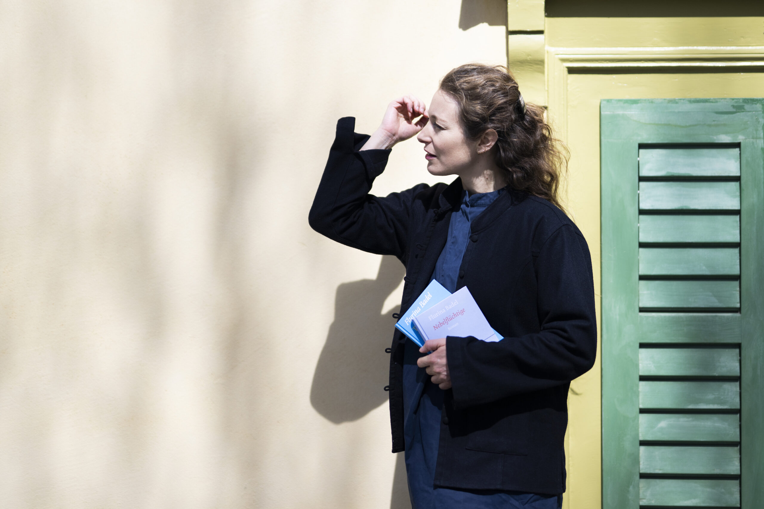 Woman in a dark blazer holds blue pamphlets while shielding her eyes with her hand, standing beside a beige wall and green shutters.