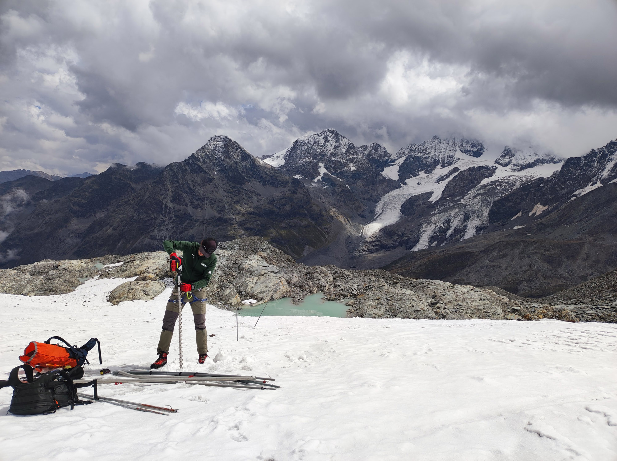 Mountaineer in a green jacket and red gloves secures gear on a snowy slope with jagged, snow-dusted peaks in the cloudy background.
