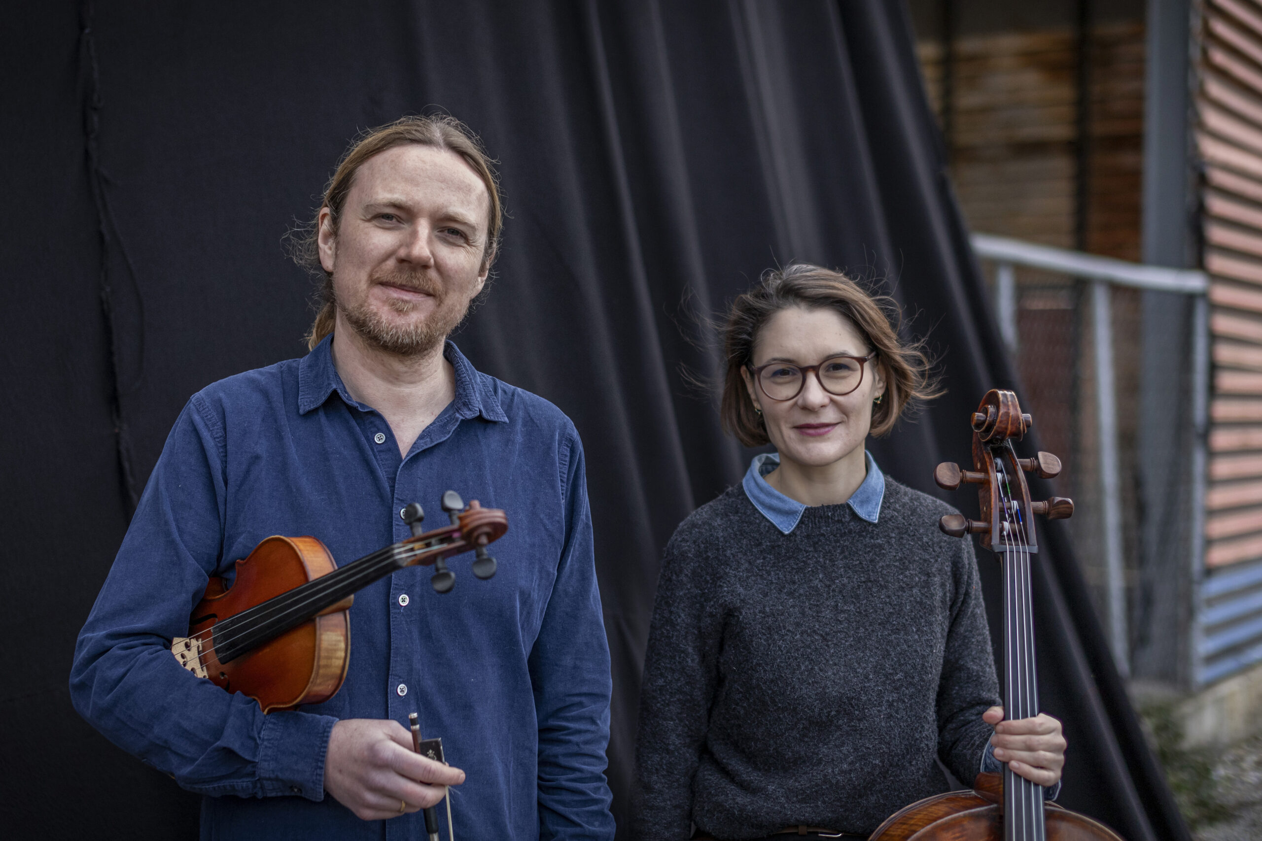 Two musicians pose with their instruments: man holding a violin in a blue shirt, woman with a cello, outdoors against a black backdrop.