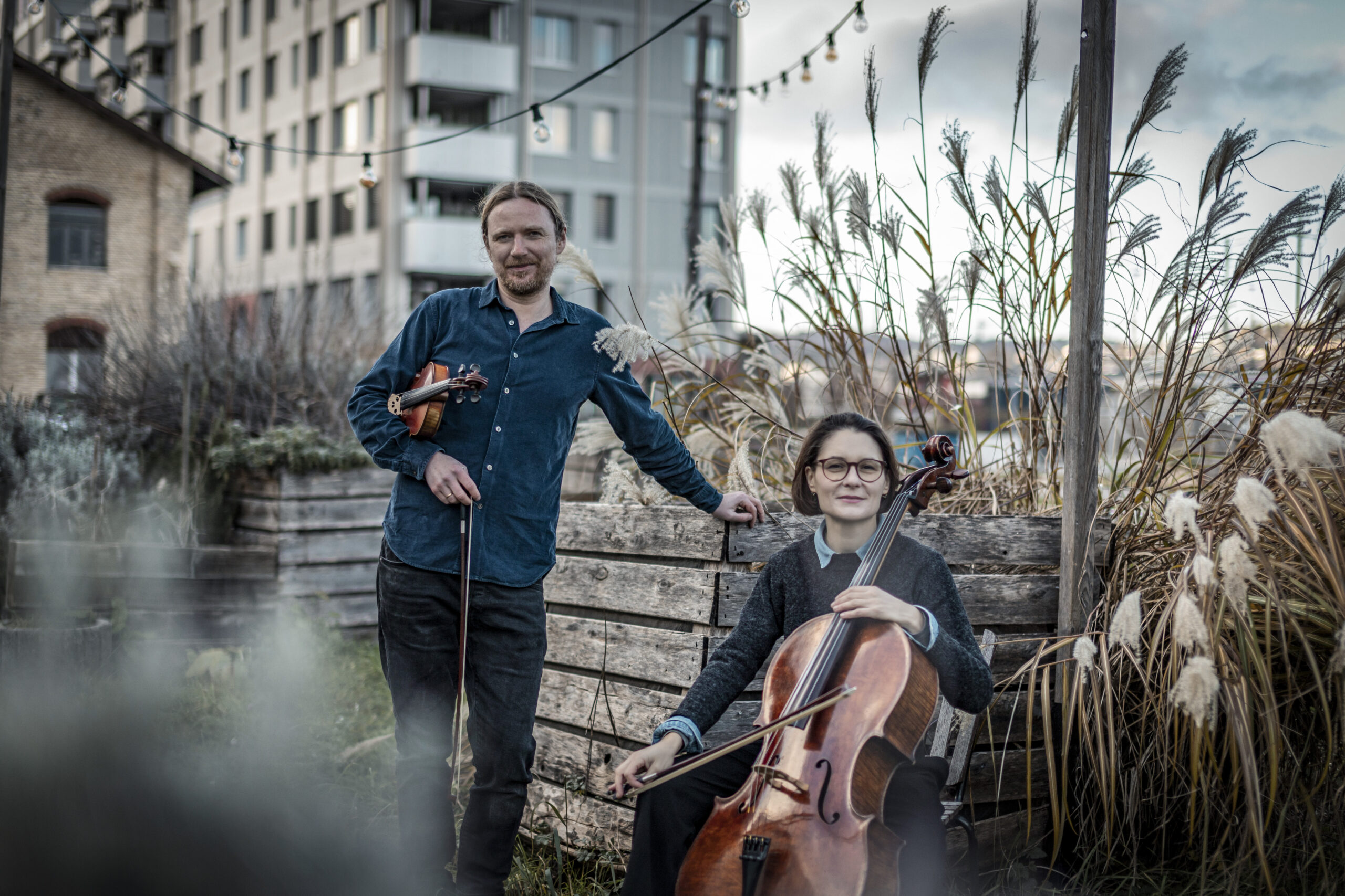 Two street musicians outdoors: a man with a violin standing beside a wooden fence, a woman seated with a cello, in a yard with tall grasses and string lights overhead.