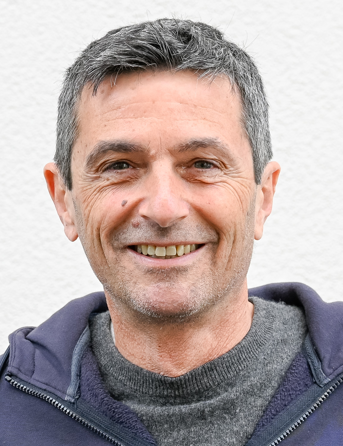 Close-up portrait of a smiling middle-aged man with short gray hair wearing a dark hoodie against a neutral background.