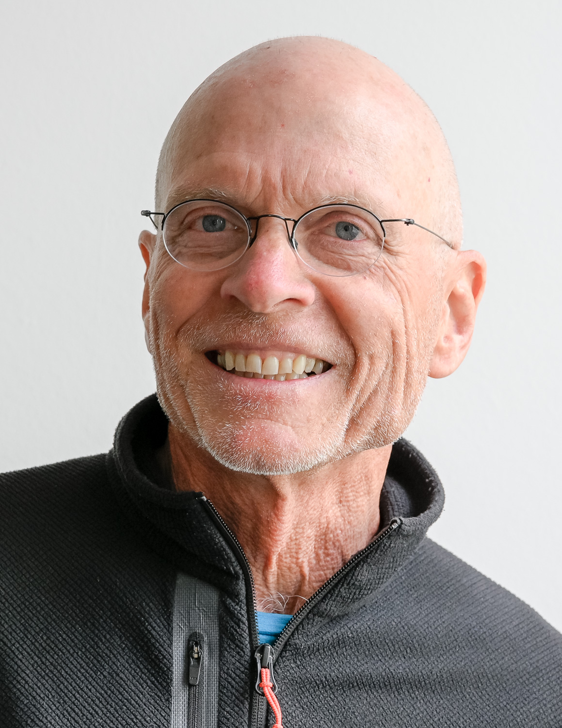 Smiling older man with round glasses and short gray beard wearing a dark zippered sweater, facing the camera.