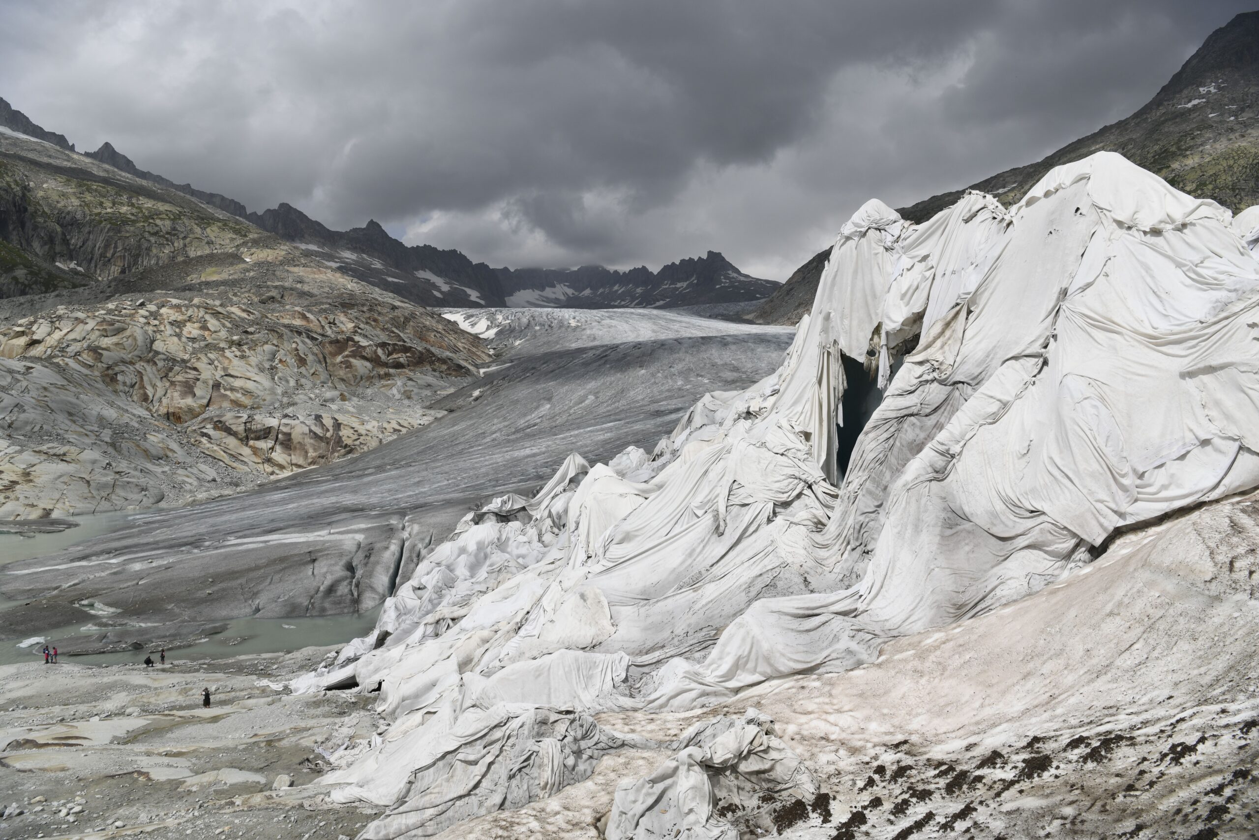Glacier valley with jagged dark mountains, a large white ice cliff in foreground, and a stormy sky; a few hikers visible in the lower left for scale.