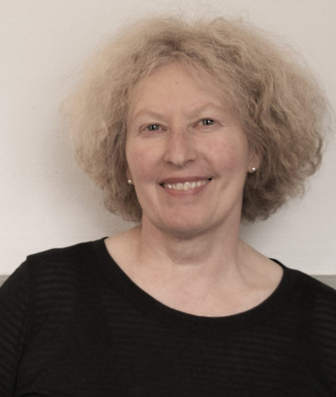 Smiling woman with curly blond hair wearing a black top, standing against a neutral wall.