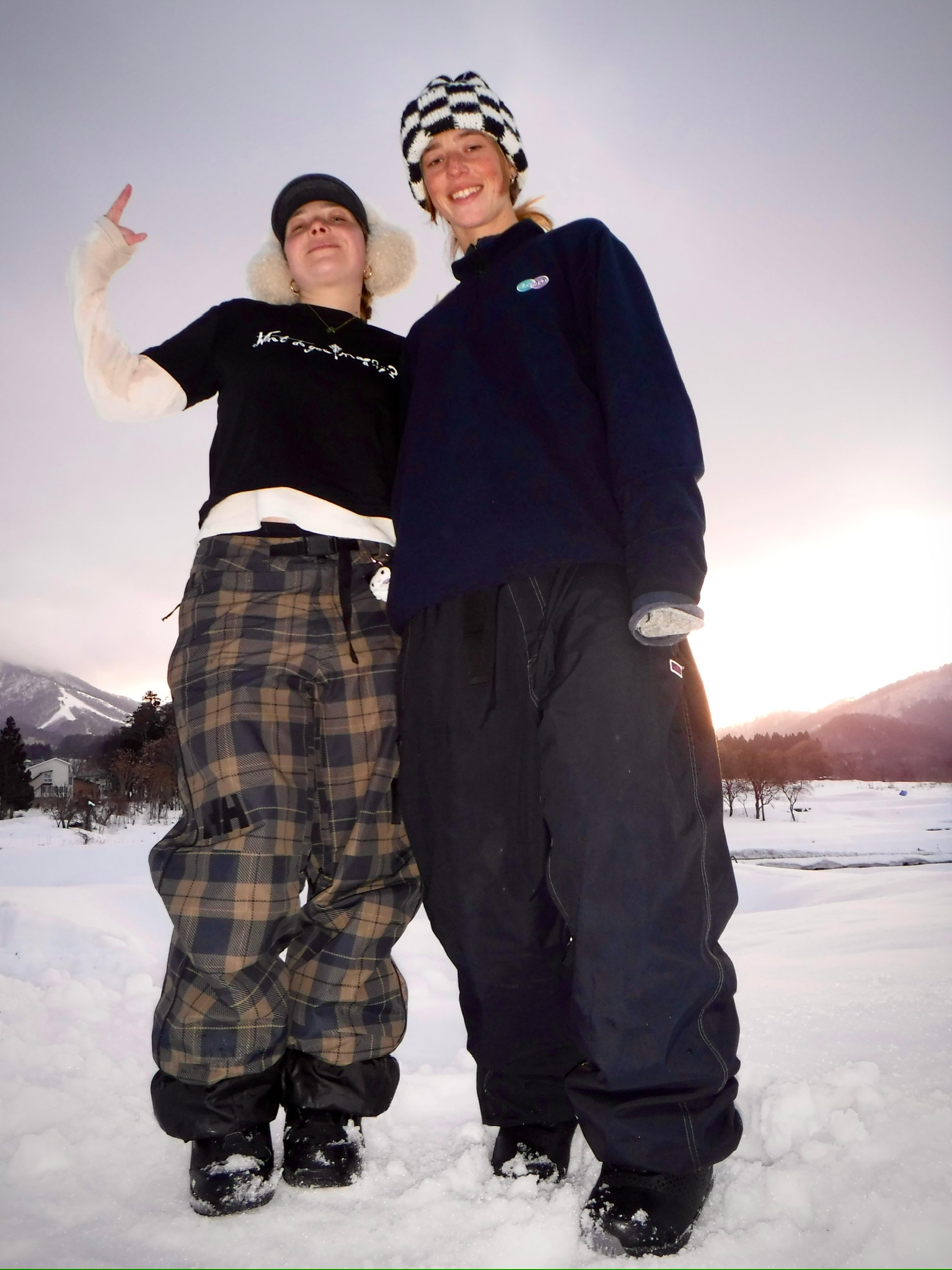 Two people stand in deep snow outdoors, wearing winter hats and jackets, smiling with a snowy landscape and mountains in the background.