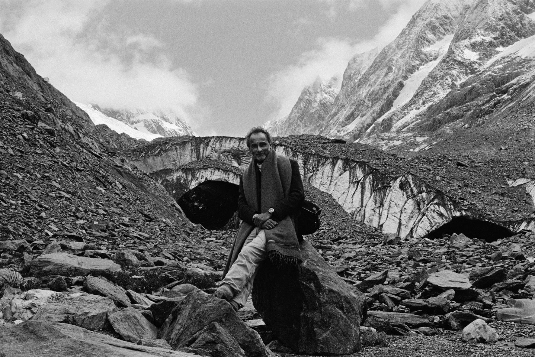Man standing on rocky mountain terrain in a scarf and coat, with snow-capped peaks in a cloudy sky behind him (black and white).