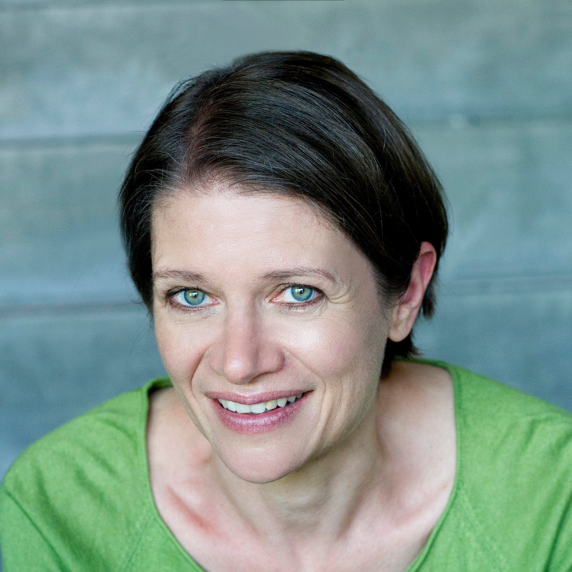 Close-up portrait of a smiling woman with blue-green eyes and dark hair, wearing a green top, cropped at the forehead.