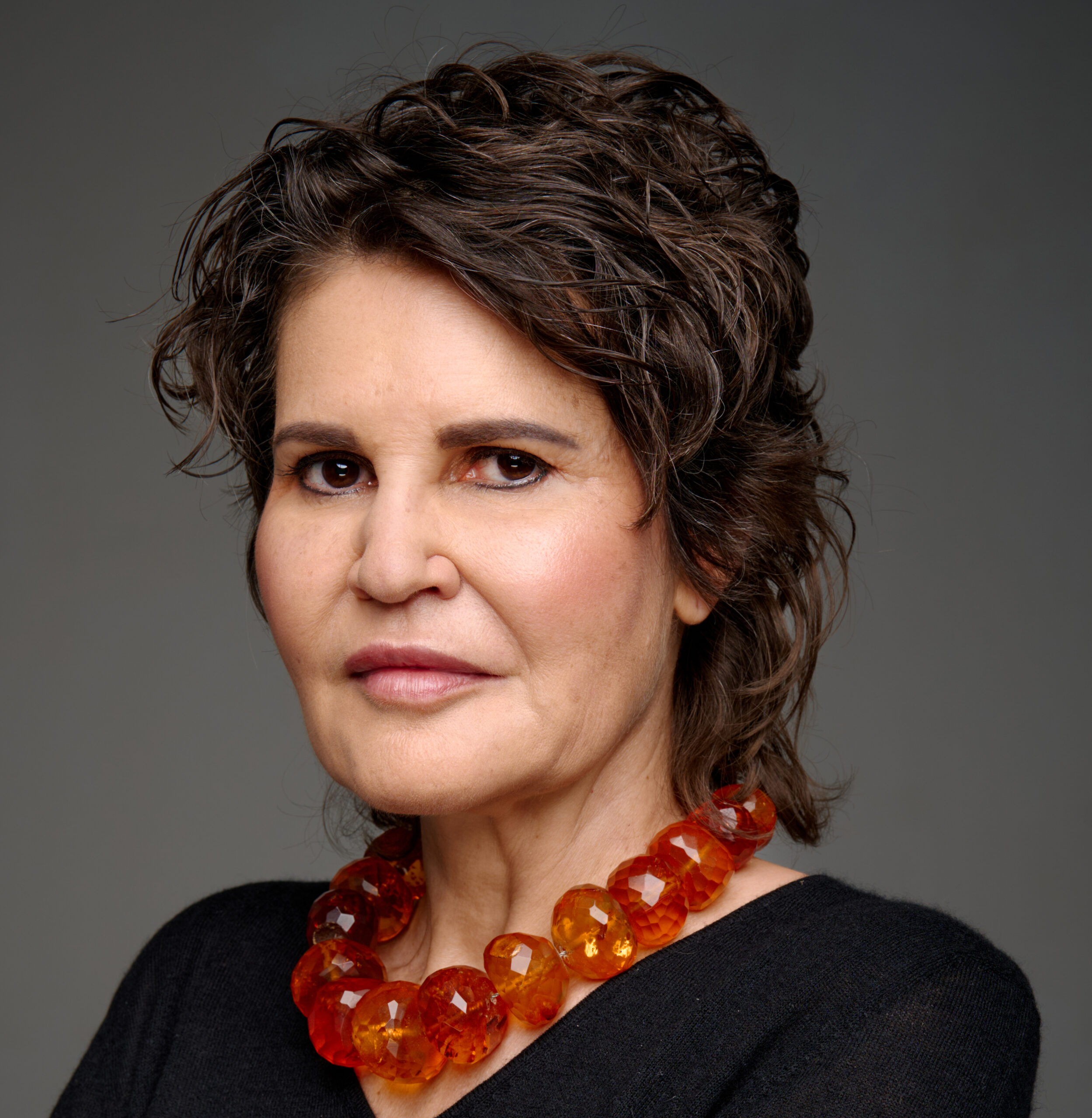 Close-up portrait of a woman with short dark hair wearing an orange beaded necklace against a gray background.