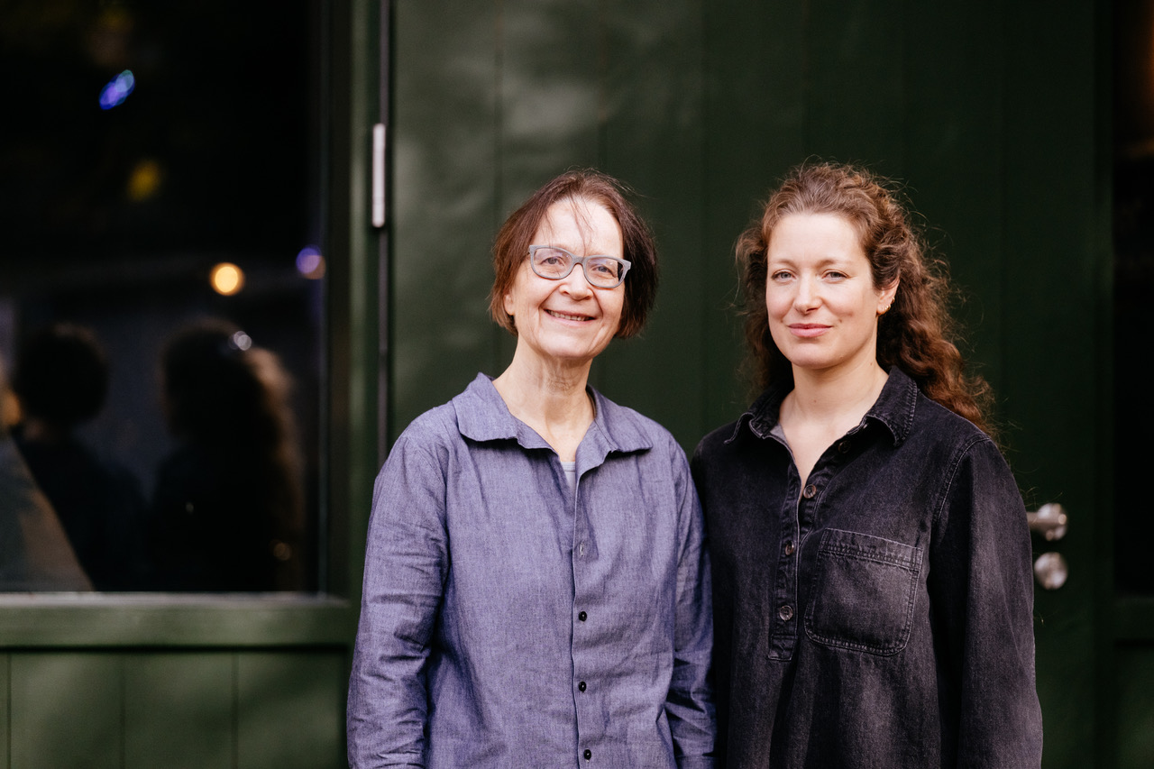 Two women standing side by side and smiling at the camera outside a dark-green door. The older woman on the left wears glasses and a light purple button-up shirt; the younger woman on the right wears a black denim shirt with her curly hair pulled back.