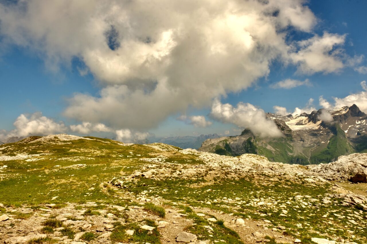 Alpine landscape with a rocky foreground, green patches, and distant snow-capped mountains under a partly cloudy blue sky.