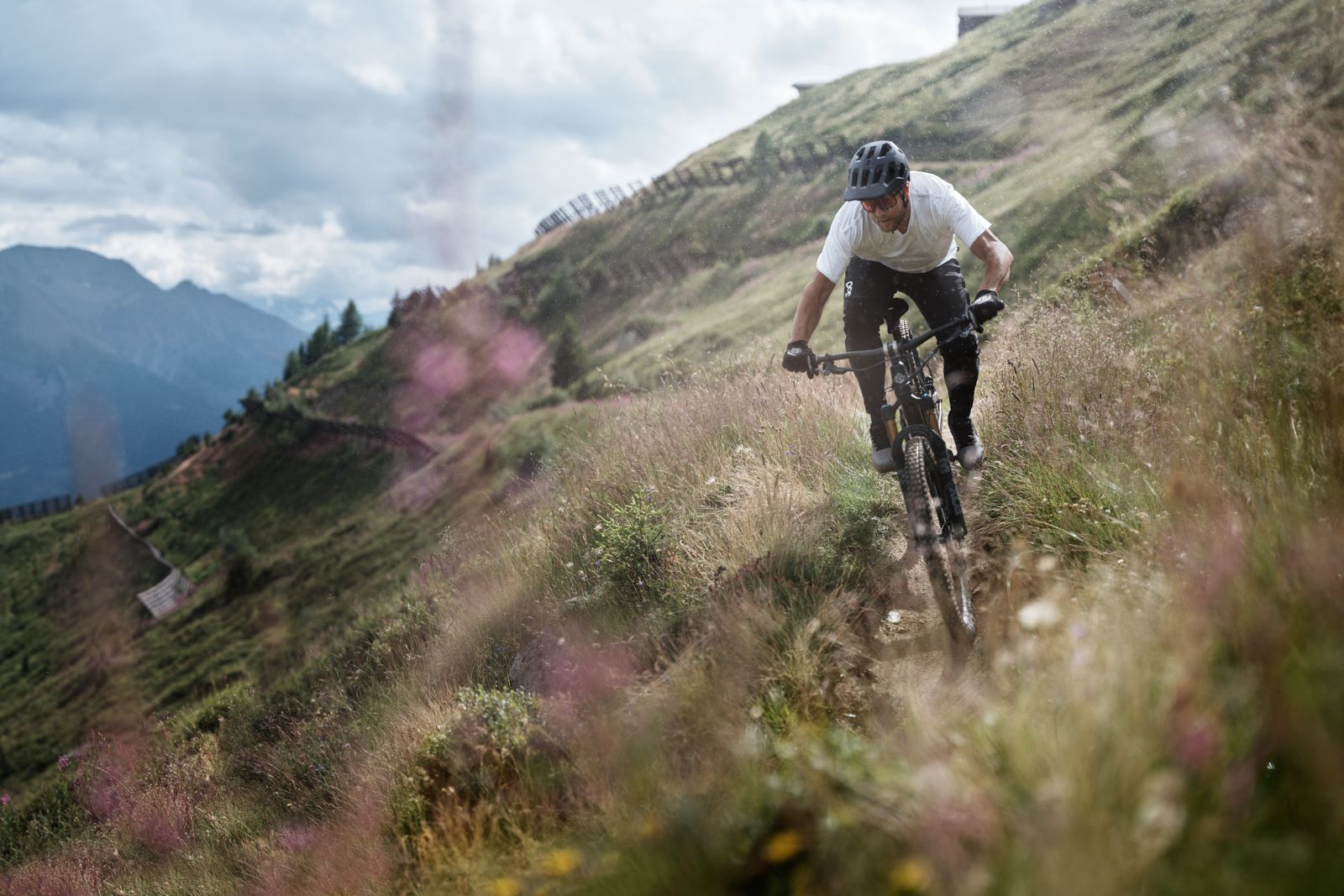 Mountain biker wearing a white shirt and helmet descends a grassy hillside on a dirt trail with mountains in the background.