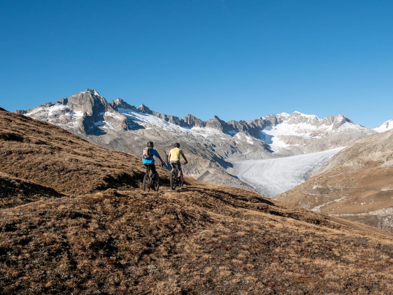 Two mountain bikers ride along a brown hillside with a snow‑capped alpine range and glacier in the distance under a clear blue sky.