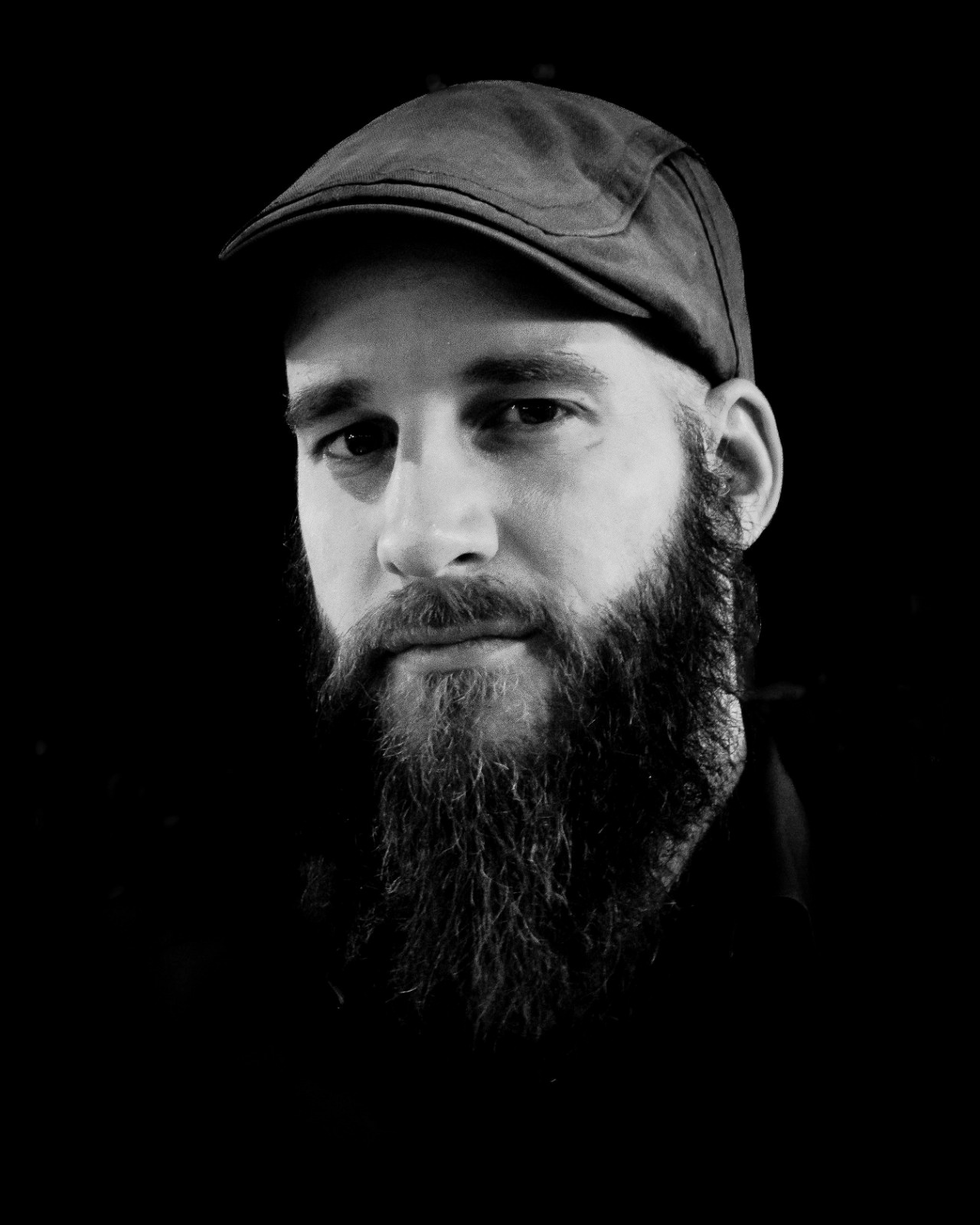 Black-and-white close-up portrait of a bearded man wearing a flat cap against a dark background, looking at the camera.