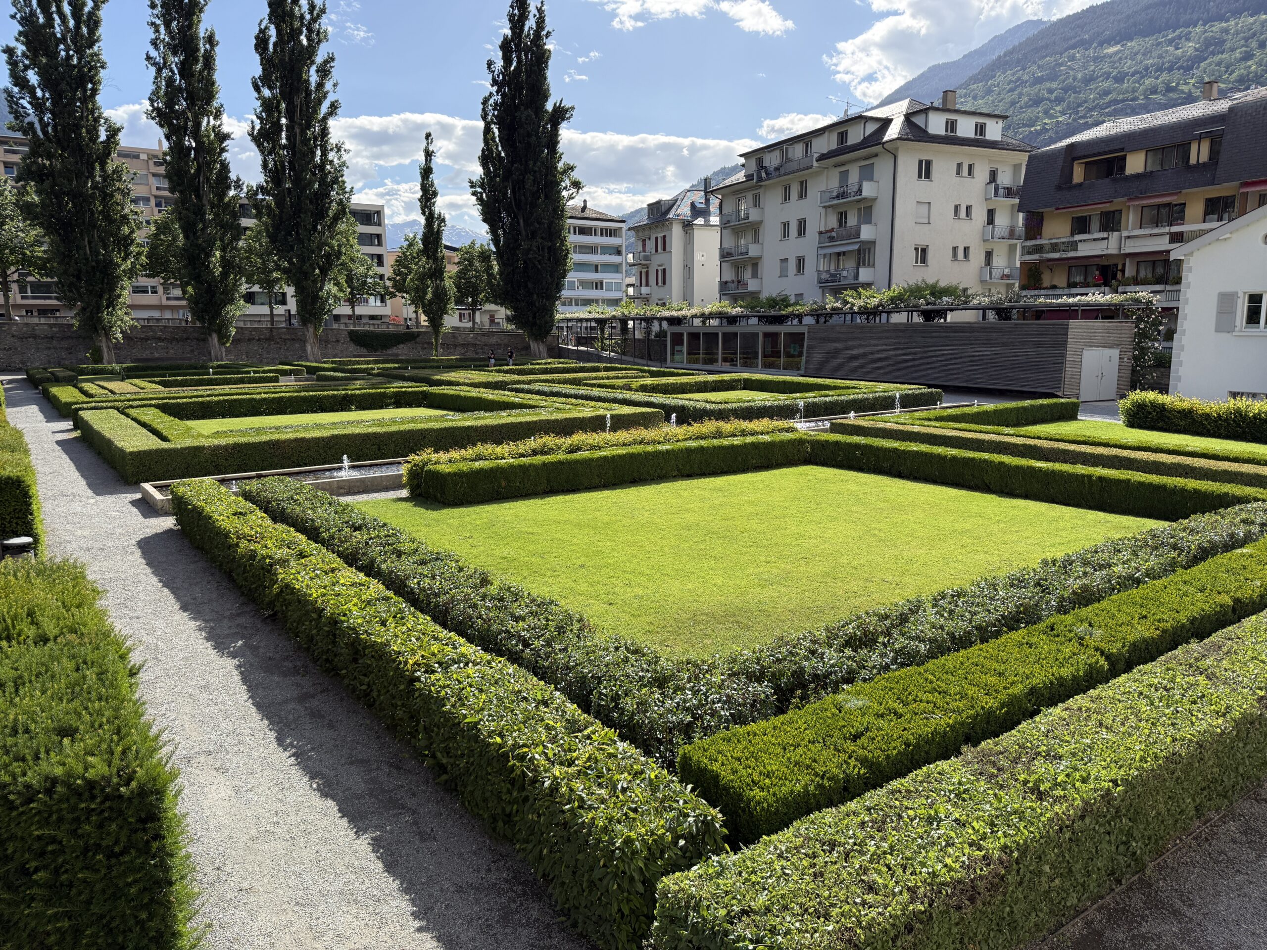 Formally clipped hedges create a geometric maze in a sunlit urban garden, with gravel paths and apartment buildings beyond.