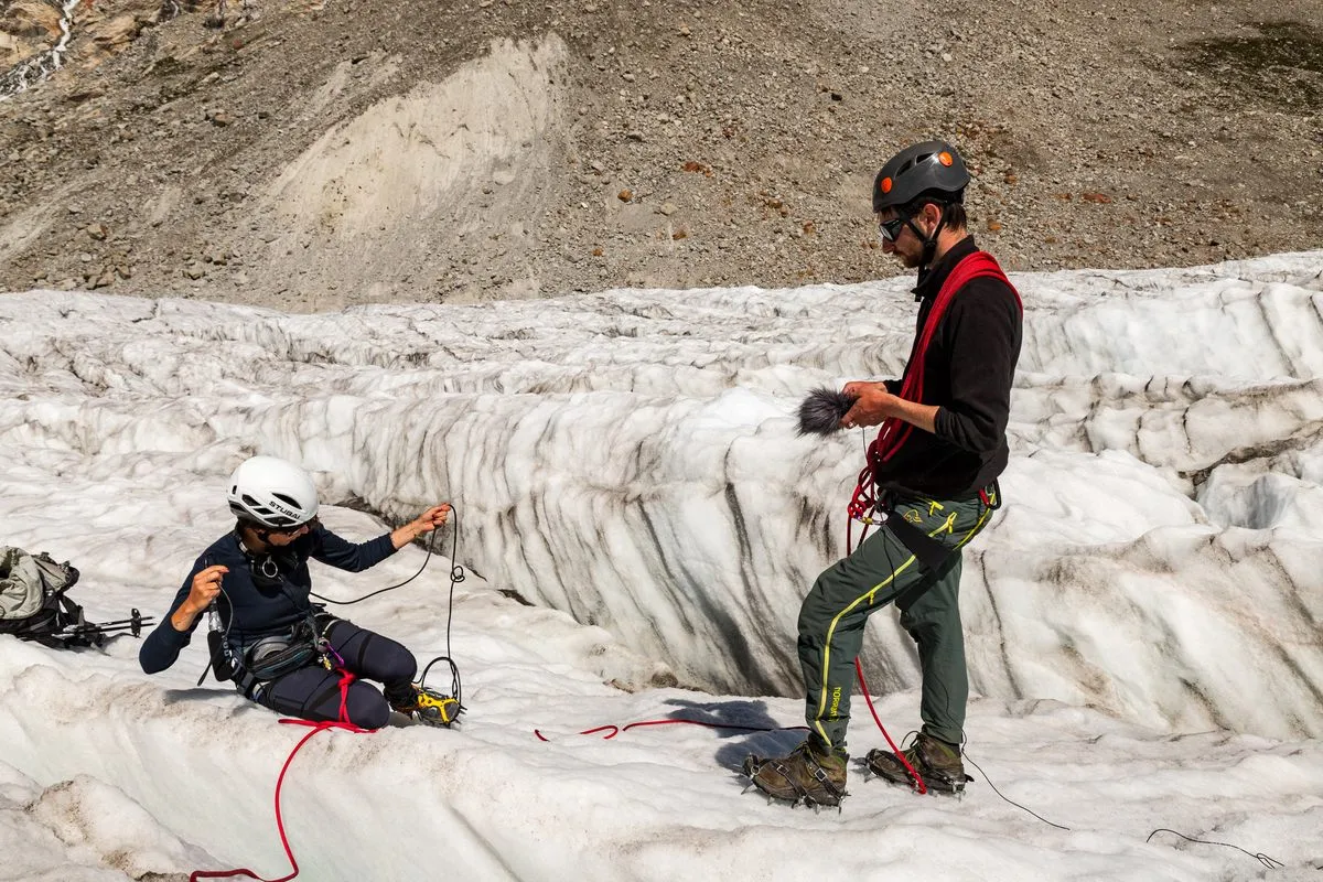 Two climbers on a glacier in helmets and harnesses; one sits and handles red rope, the other stands with a furry microphone windscreen and gear behind.
