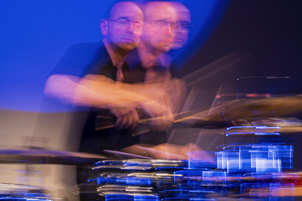 Blurred motion of a drummer playing a drum kit on stage under blue lighting during a live performance