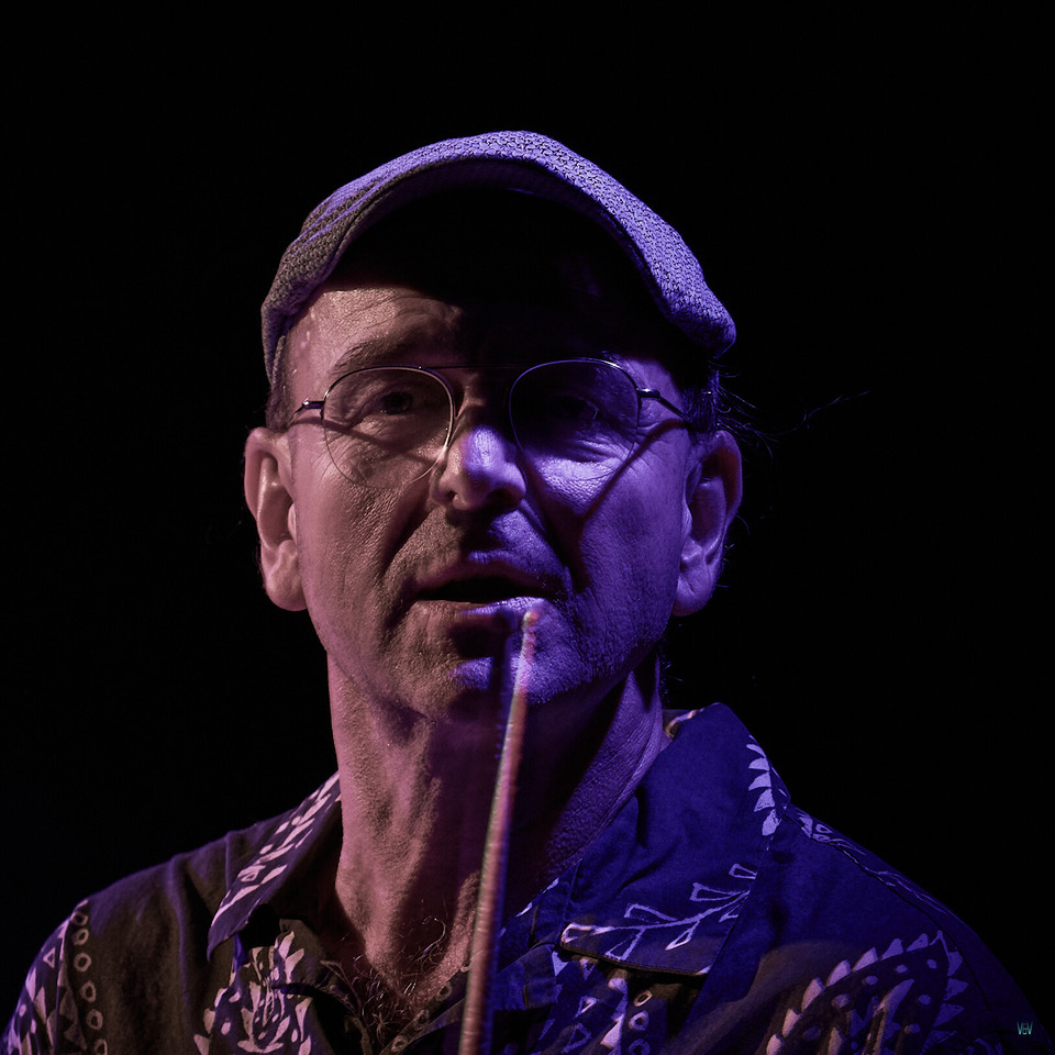 Older man wearing a flat cap and glasses, smoking a cigarette under purple stage lighting on a dark stage.