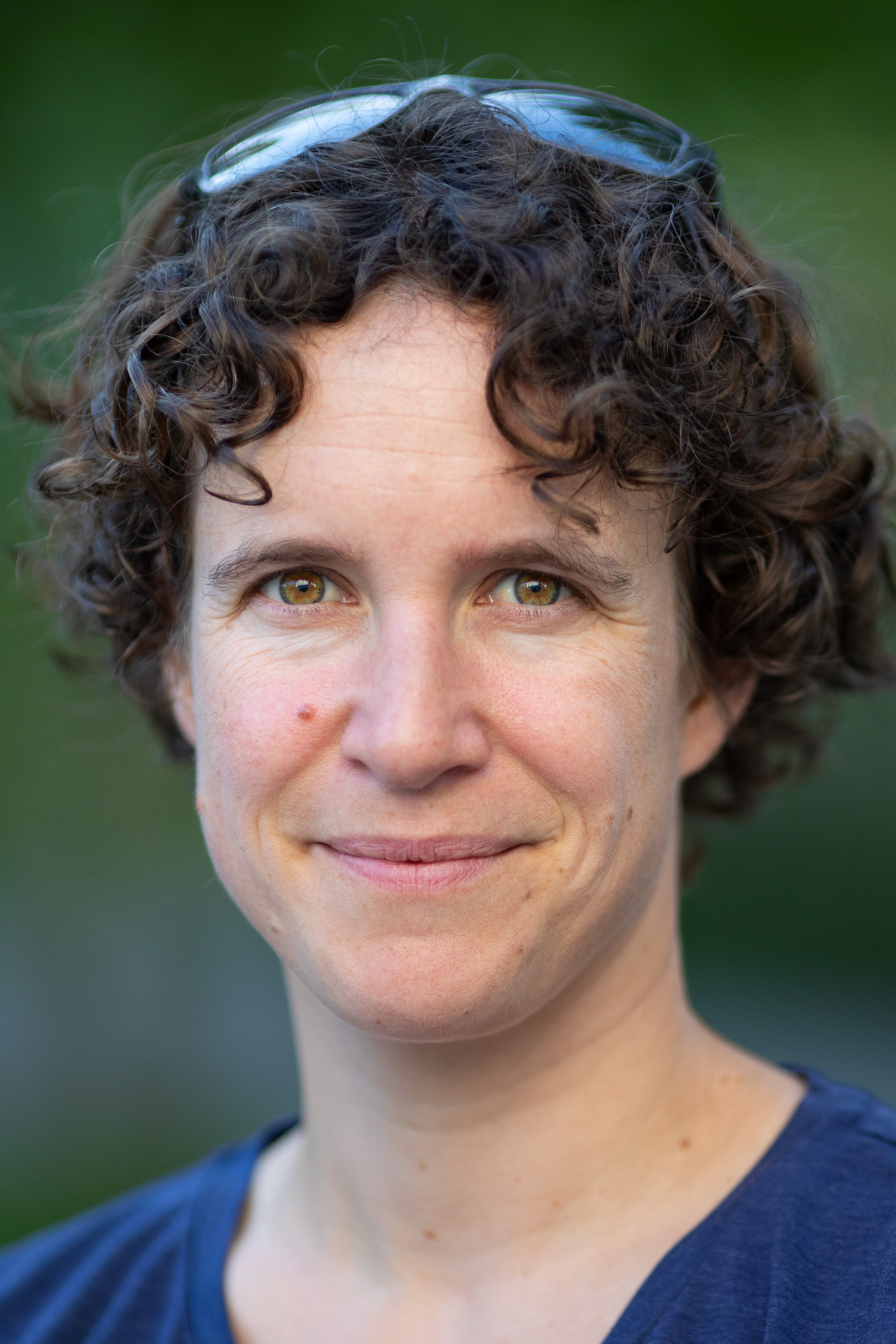 Close-up portrait of a smiling woman with short curly hair, wearing sunglasses on her head and a blue shirt, outdoors.