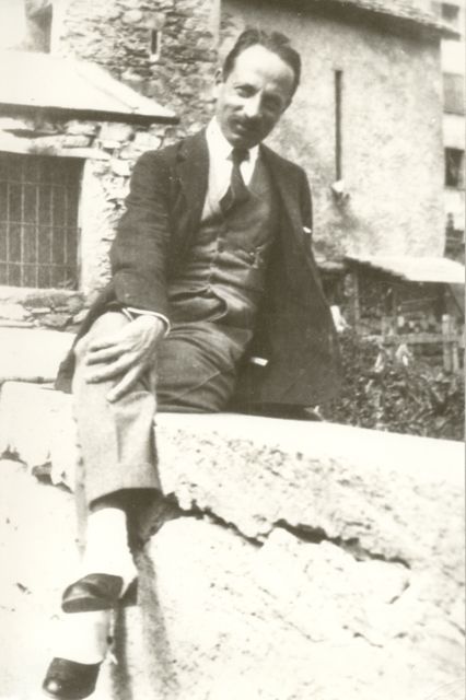 Man in a suit sits on a rough stone wall outdoors, legs crossed, posing for the camera beside a rustic building.