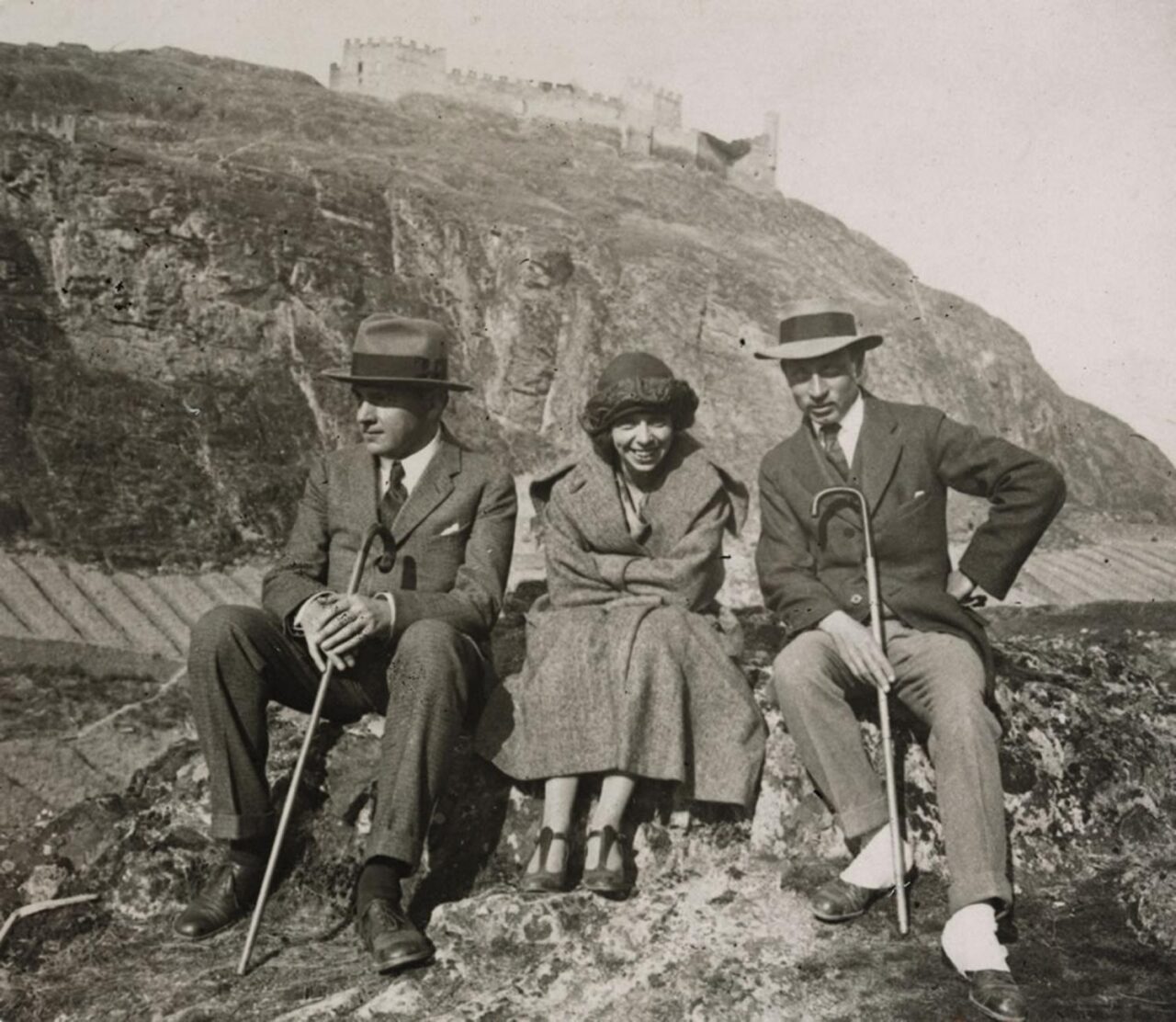 Three people sit on a rocky ledge with a castle on a hill in the background: two men in suits and hats with canes flank a smiling woman in a coat and hat.