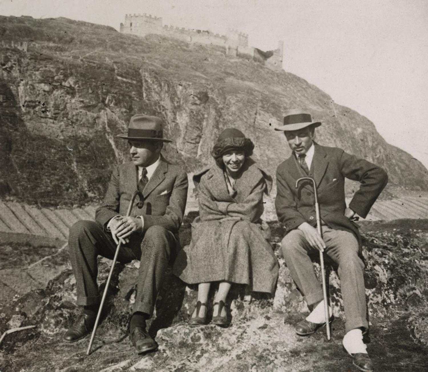 Three people sit on a rocky ledge with a castle on a hill in the background: two men in suits and hats with canes flank a smiling woman in a coat and hat.
