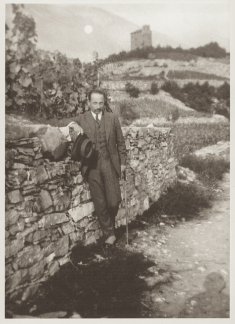 Gentleman in a suit leaning on a stone wall with a cane and hat, in a vineyard with a hilltop castle in the background.