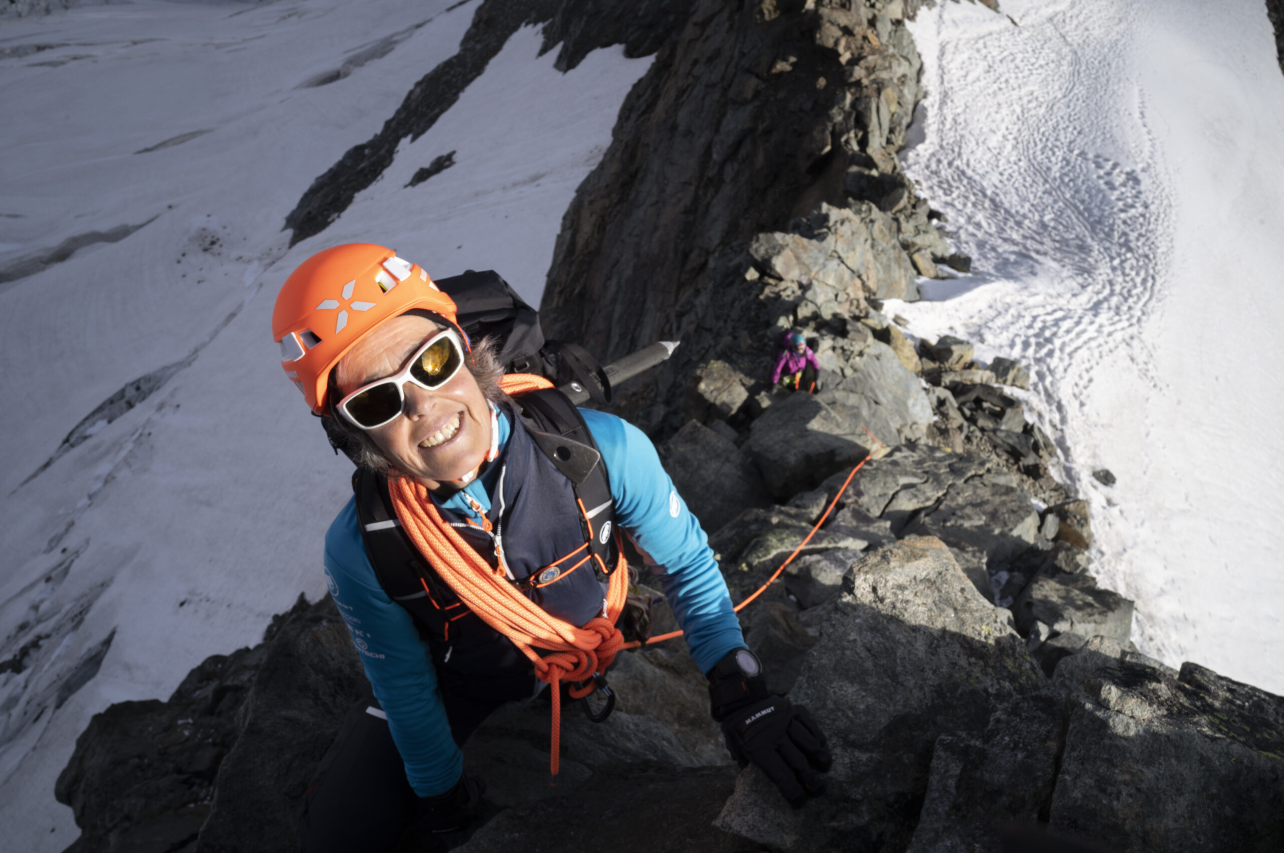 Mountaineer wearing an orange helmet and blue jacket climbing rocky ridge above snow and ice, secured by ropes.