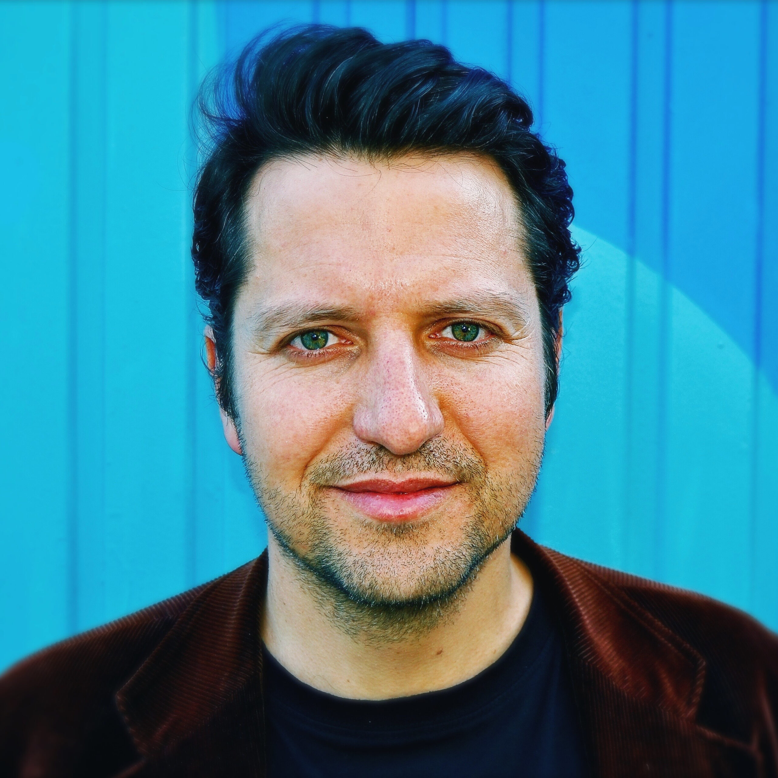 Close-up of a man's face from the nose up, with green eyes and dark hair against a blue background on a turquoise wall.