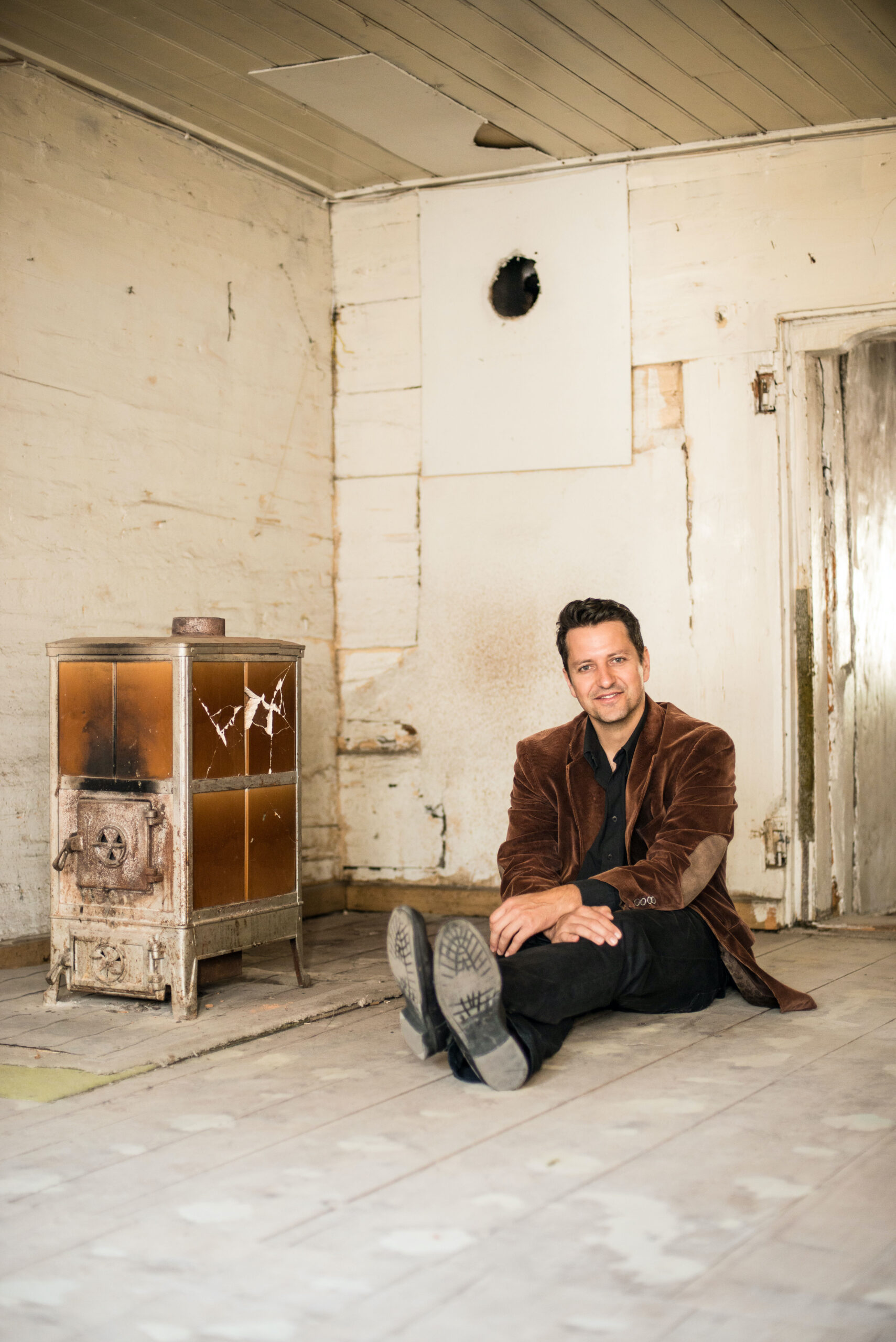 Smiling man in a brown blazer sits on a dusty wooden floor in a dilapidated room with an old rusty stove nearby.