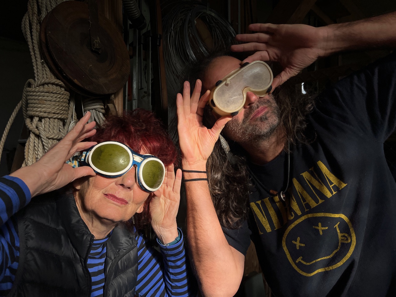 Two adults wearing oversized protective goggles pose playfully in a workshop filled with ropes and coils in the background.