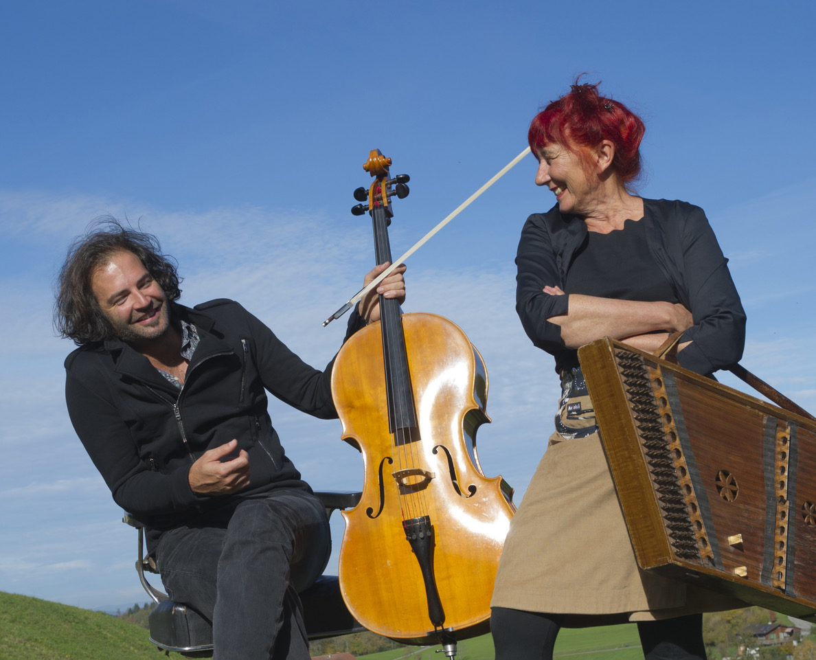 Two street musicians outdoors: a man with a cello and a woman with a hurdy-gurdy, smiling at each other under a blue sky.
