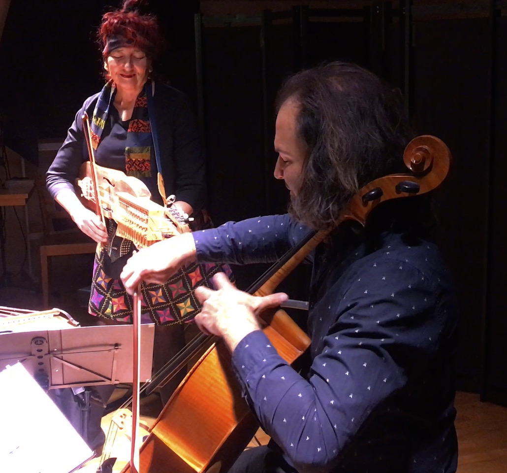 Two musicians perform on a stage: a woman plays a stringed instrument beside a seated cellist who is bowing.