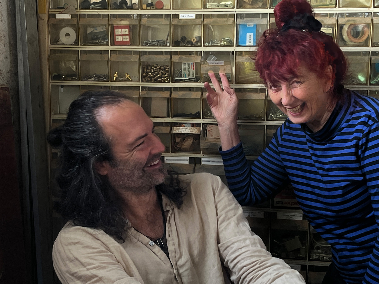 Two people laughing and high-fiving in front of a wall of small labeled drawers in a workshop/gallery of hardware.
