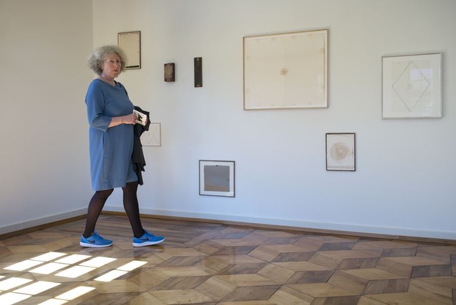 Woman in a blue dress walks through a gallery, looking at framed artworks on a pale wall.
