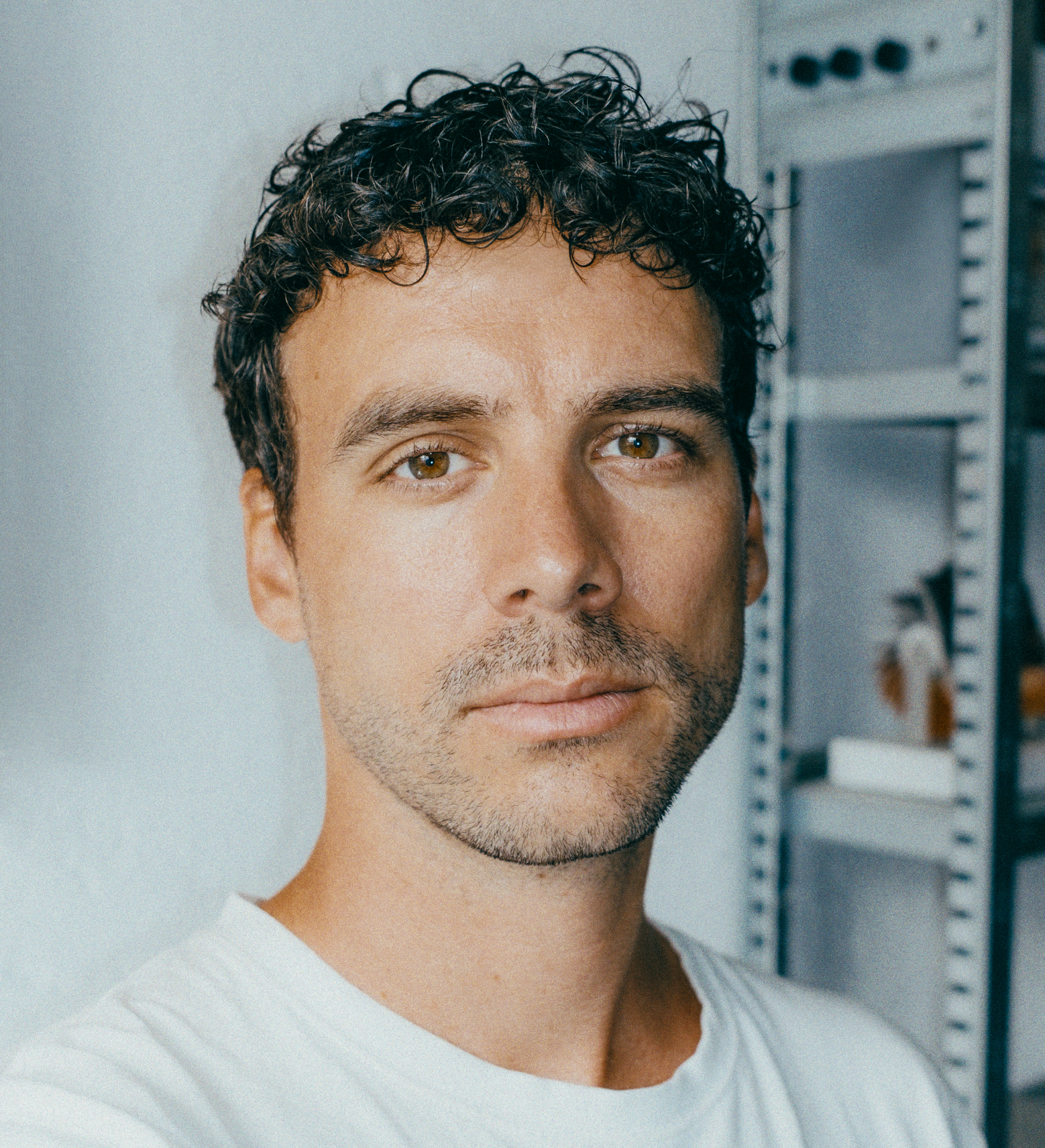 Close-up of a young man's face (eyes and mouth) wearing a white shirt, with a blurred workshop shelf in the background.