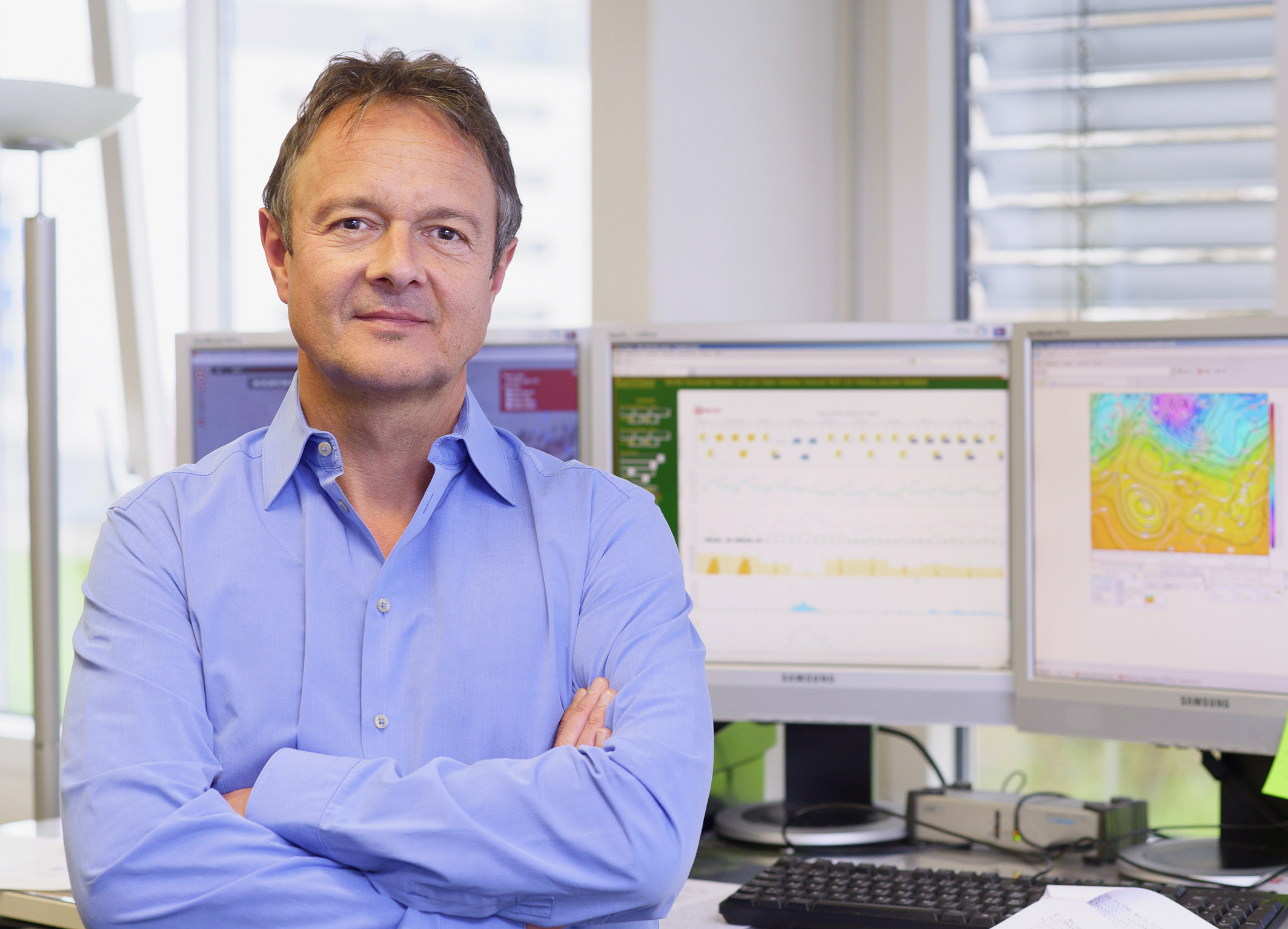 Man in a light blue button-down shirt stands with arms crossed in front of computer monitors in an office setting, looking at the camera.