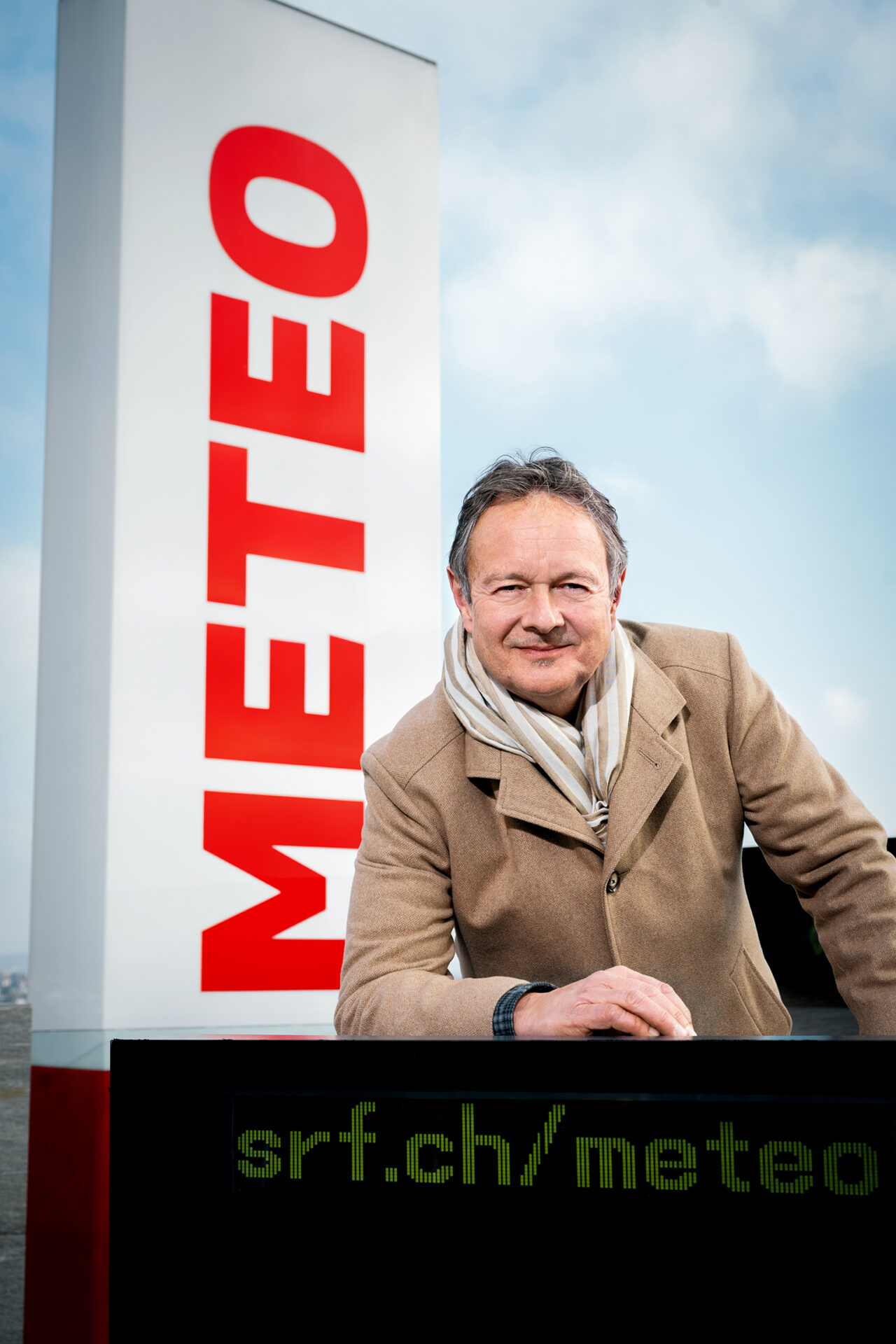 Smiling man in a beige coat and striped scarf leaning on a black display panel beside a tall white sign with red vertical lettering outdoors.] ,