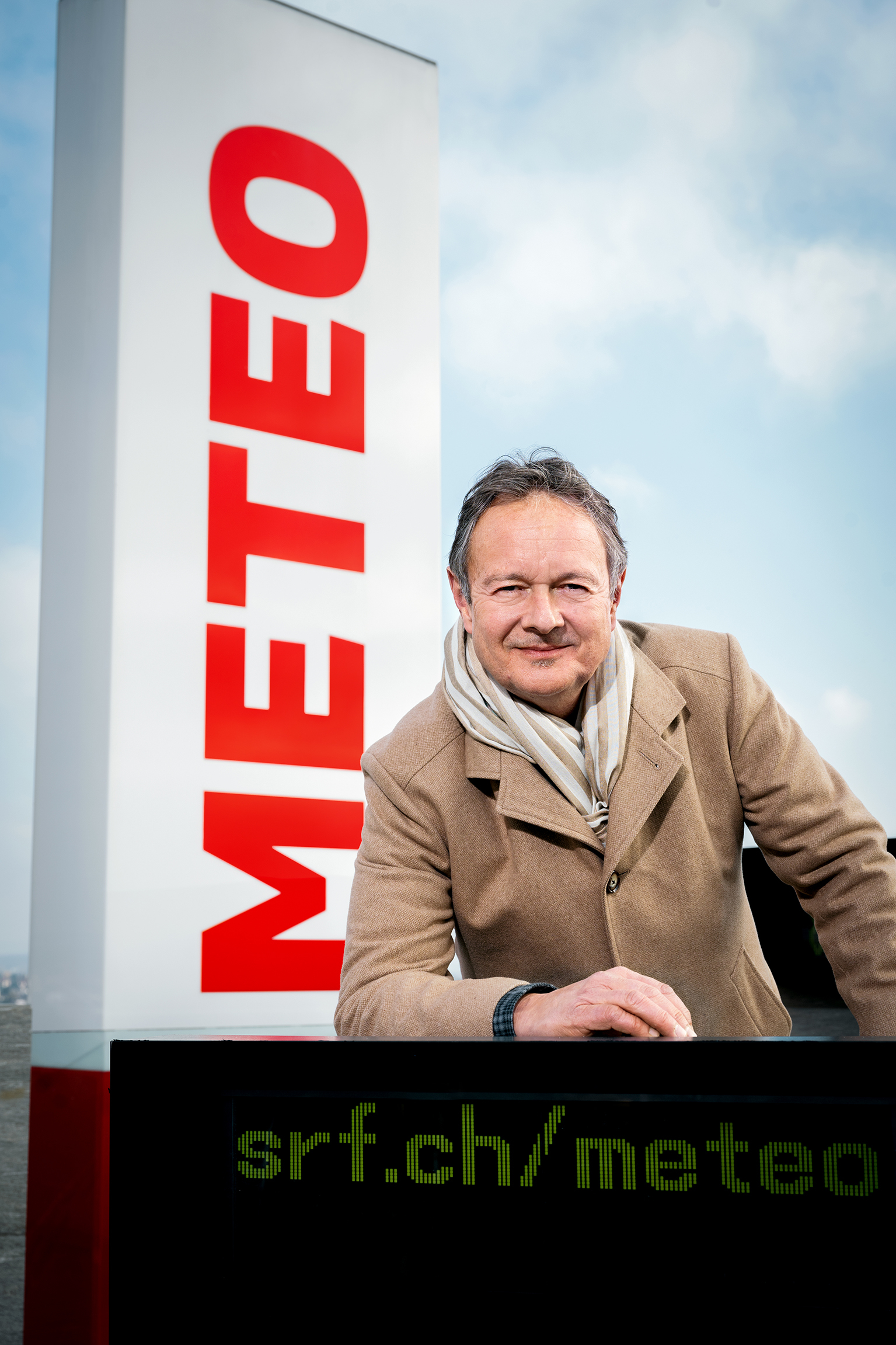 Smiling man in a beige coat and striped scarf leaning on a black display panel beside a tall white sign with red vertical lettering outdoors.] ,