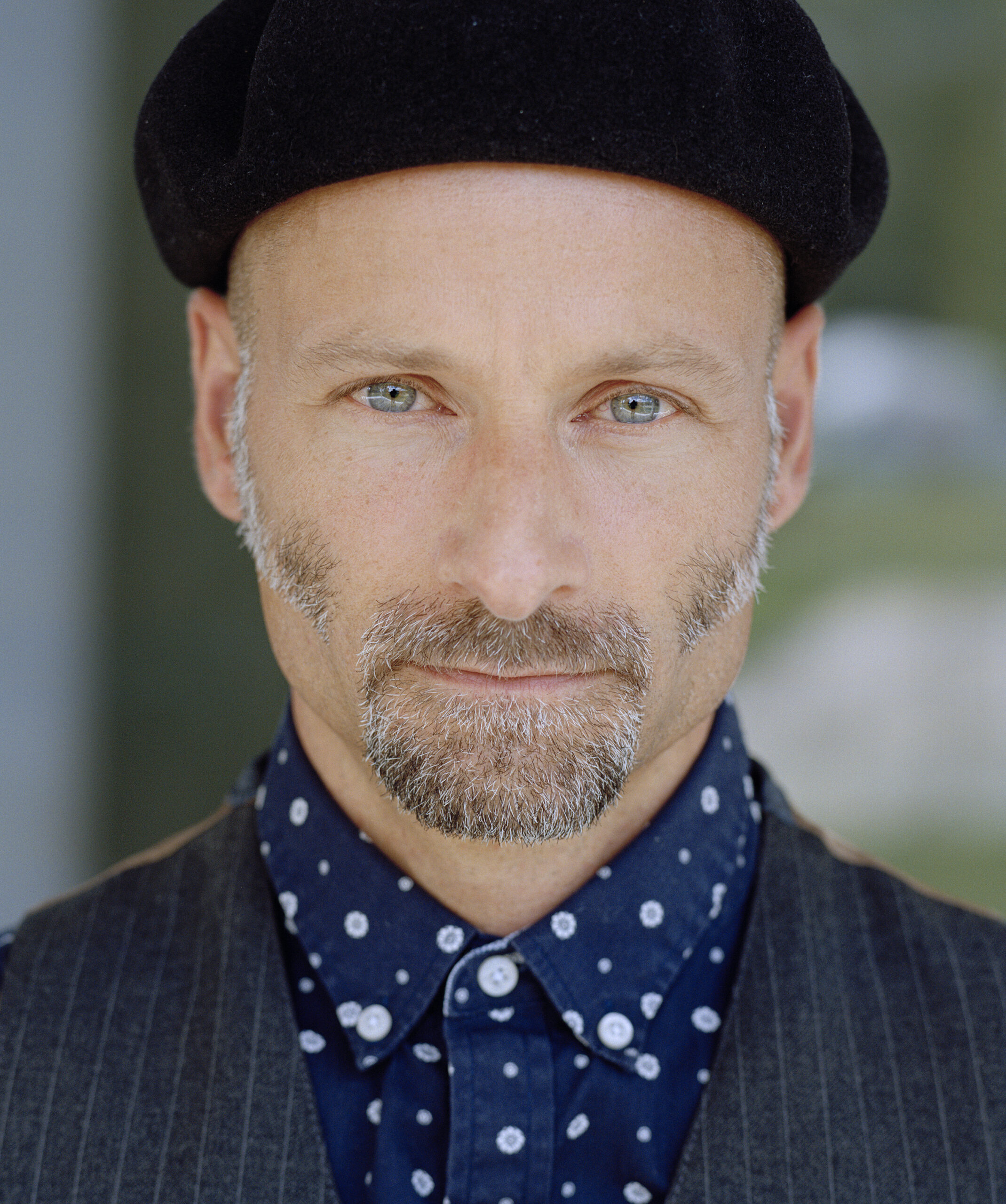 Close-up portrait of a man wearing a black beret, with light stubble and a navy polka-dot shirt outdoors.