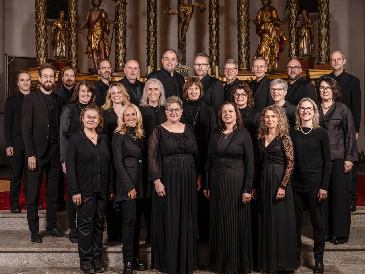 A large choir of about twenty people in black formal attire posing on church steps with ornate statues and columns behind them.