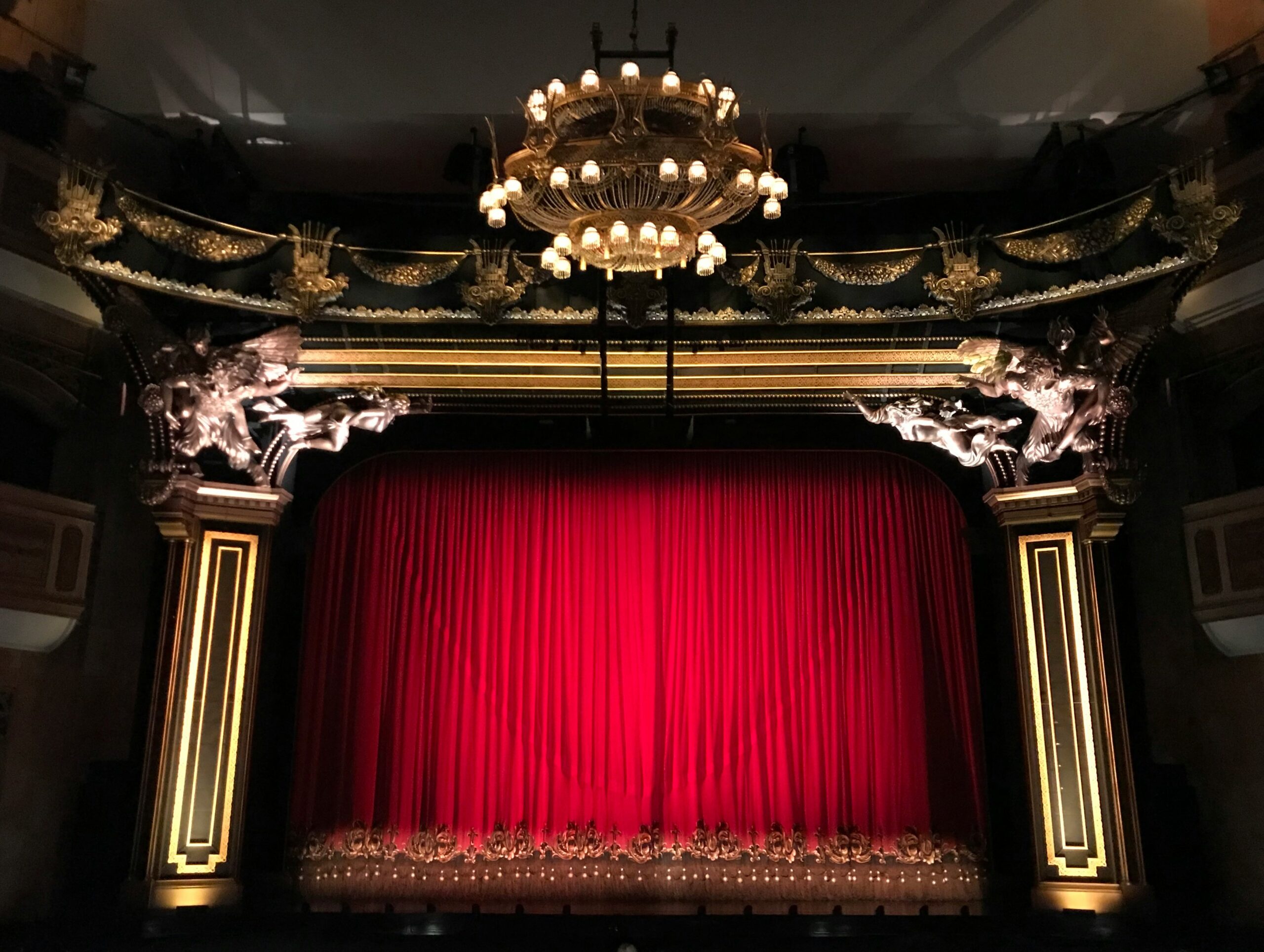 Ornate theater stage with a red velvet curtain and ornate gold trim along the proscenium and balcony railings, chandeliers overhead.