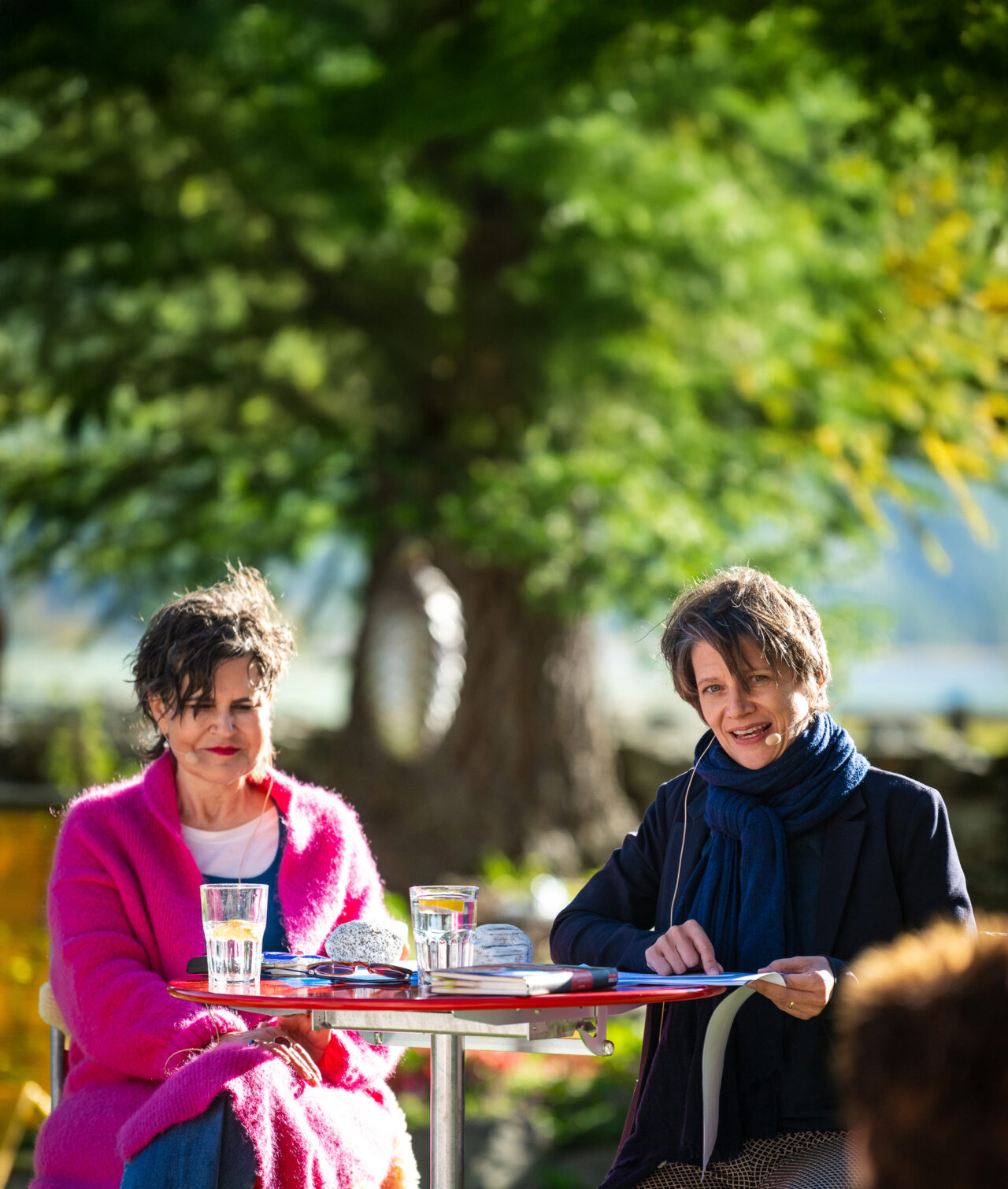 Two women sit at a red outdoor table with drinks, papers, and microphones, smiling in a sunny garden setting.