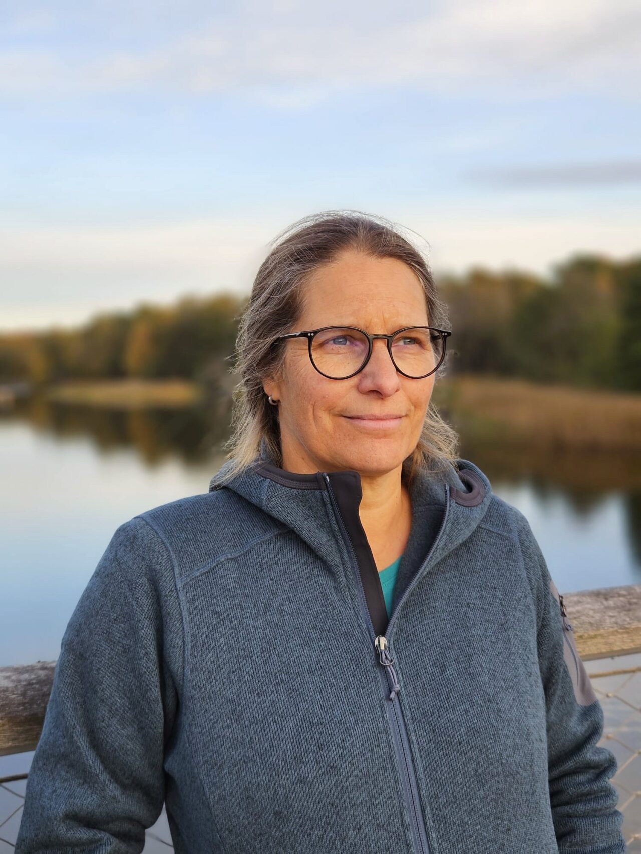 Portrait of a smiling woman with glasses, standing outdoors by a lake with trees in the distance, wearing a blue zip-up jacket.
